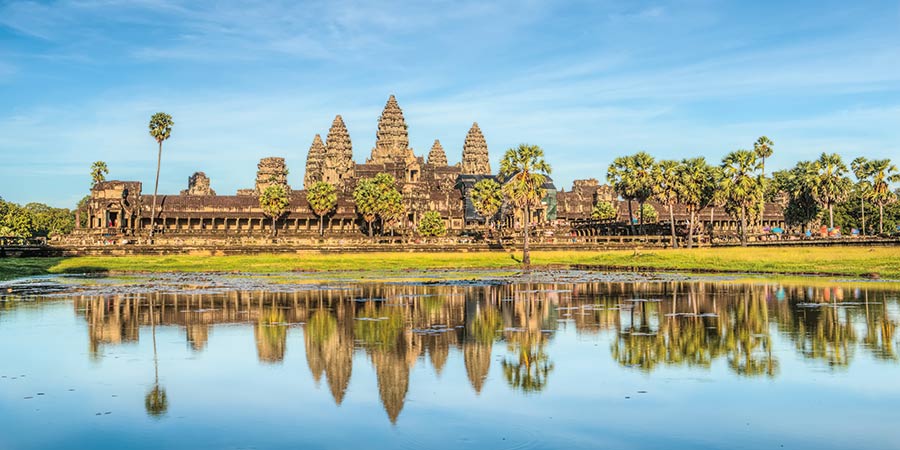 View of Angkor Wat, a large ancient temple complex in Cambodia. The temple's towering stone spires rise above the surrounding trees, with intricate carvings visible on the walls. The scene is bathed in warm sunlight, and blue skies all reflecting off a nearby water in the foreground. 