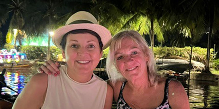 Two women smiling together in front of a tropical river at night, surrounded by palm trees, boats and lighting. 